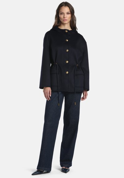 Cappotto in lana blu navy con cappuccio, con bottoni dorati, coulisse in vita e due tasche frontali. Abbinato a jeans a gamba larga e tacchi.