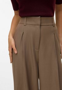 Pantalones marrones ajustados con cintura alta, pliegues y bolsillos laterales. La tela parece suave con un ligero brillo, complementada por una blusa burdeos.