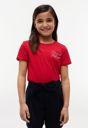 Smilende pige med langt brunt hår iført en rød Tommy Hilfiger t-shirt og marineblå shorts bundet med en sløjfe.
