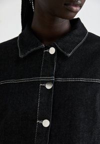 Black denim jacket featuring a classic collar, white contrast stitching, and silver snap buttons along the front placket.