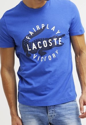 Mężczyzna noszący niebieską koszulkę z czarną grafiką krokodyla i białym tekstem z napisem „Fairplay Lacoste Victory.”