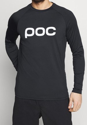 Mężczyzna ubrany w czarną koszulkę z długim rękawem z białym logo "poc" na klatce piersiowej oraz czarne spodnie.