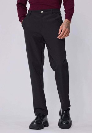 Homme portant un pantalon noir droit, des chaussures noires épaisses et un haut bordeaux à manches longues, debout les mains dans les poches.