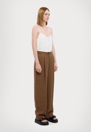 White camisole top with thin straps paired with loose, brown, pleated trousers and chunky black sandals, against a plain background.