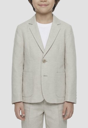 Enfant portant un blazer beige clair sur une chemise blanche, debout devant un fond gris uni.