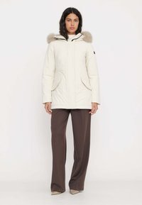 Parka bianca con cappuccio foderato in pelliccia, chiusura frontale con zip e due grandi tasche. Abbinata a pantaloni marroni a gamba larga e scarpe beige.