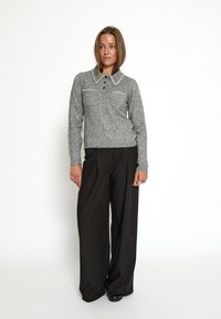 Donna in piedi con indosso un maglione grigio lavorato a maglia con bottoni e colletto, e pantaloni neri a gamba larga su sfondo bianco.