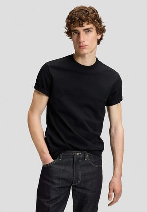 Junger Mann mit lockigem Haar, der ein schwarzes T-Shirt mit kurzen Ärmeln und dunkle Jeans trägt, steht mit einer Hand in der Tasche.