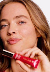 Czerwony produkt do ust NUXE z błyszczącym wykończeniem i srebrnymi akcentami. Modelka z falującymi włosami trzyma tubkę w miękkim rozmyciu, prezentując gładką skórę.