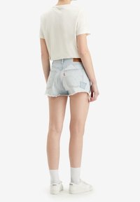 Camiseta blanca de corte recto y pantalones cortos de mezclilla azul claro con dobladillo deshilachado. Combinados con calcetines y zapatos blancos. Destaca la marca Levi's en la banda de la cintura.
