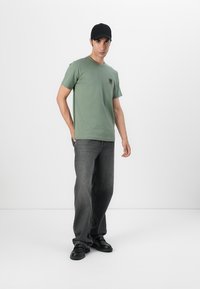 Tricou din bumbac de culoare verde măsliniu cu un mic logo pe piept, asortat cu blugi gri închis și încălțăminte neagră, pe un fundal alb.