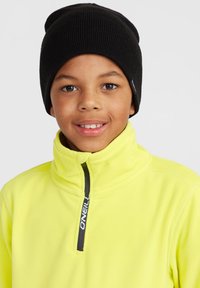 Bonnet en tricot noir et veste en polaire jaune vif avec fermeture éclair, arborant un détail de logo noir. Texture lisse et design chaud.