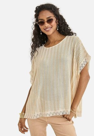 Mujer con top de punto beige claro con borde de encaje, pantalones beige, gafas de sol estilo cat-eye y joyas doradas, sonriendo frente a un fondo blanco.