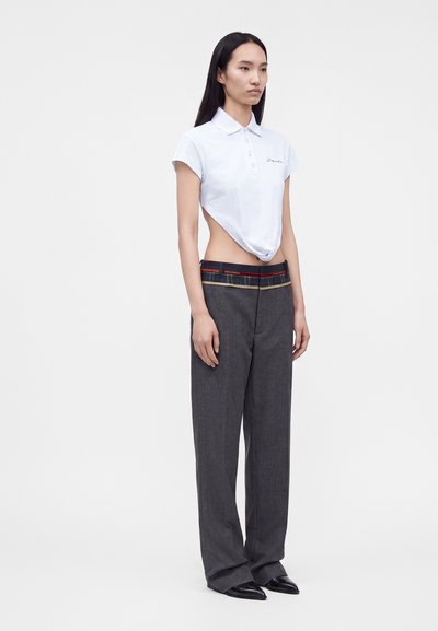 Femme portant un polo blanc court associé à un pantalon gris taille haute à jambes larges et des chaussures noires pointues, debout devant un fond uni.