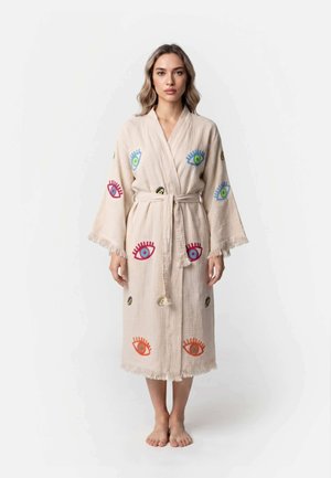 Vrouw staat blootsvoets en draagt een beige badjas met kleurrijke geborduurde oogpatronen en een gebonden ceintuur tegen een effen witte achtergrond.