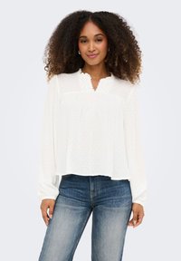 Γυναίκα με σγουρά μαλλιά που φοράει λευκή μπλούζα με μακριά μανίκια και δαντελένιες λεπτομέρειες, καθώς και μπλε τζιν, όρθια μπροστά σε ένα απλό φόντο.