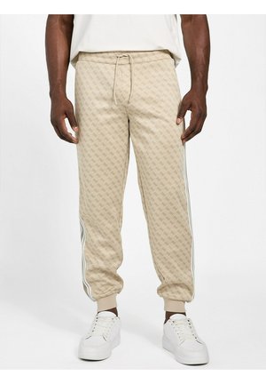 Uomo che indossa pantaloni jogger beige con fantasia e bande laterali e sneaker bianche, in piedi davanti a uno sfondo bianco semplice.