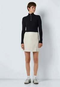 Blusa preta de manga longa com fecho éclair combinada com uma saia curta bege clara até ao joelho, com uma fenda frontal, usada com meias brancas e sapatilhas cinzas.