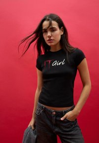 Młoda kobieta z ciemnymi włosami, ubrana w czarny T-shirt z napisem "IT Girl" i dżinsy, stoi na tle czerwonym, trzymając w jednej ręce szarą kurtkę.