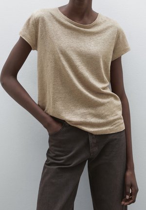 Dark-skinned person wearing a beige short-sleeve top and dark brown pants, with one hand in pocket, standing against a plain light wall.
