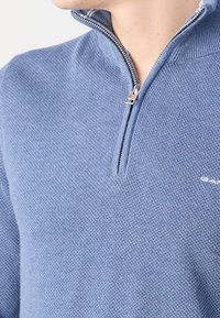 Hellblaues Reißverschlussoberteil aus strukturiertem Strickstoff, mit hohem Kragen und silbernem Reißverschluss, sowie einem kleinen Logo auf der Brust.