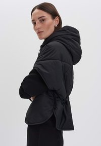 Veste noire matelassée avec capuche, présentant un design matelassé, une taille froncée et des manches courtes. Texture lisse et coupe décontractée.