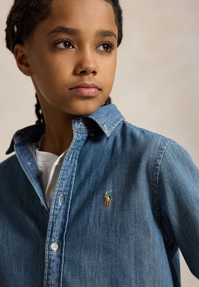 Jeune personne aux cheveux tressés portant une chemise en jean bleue par-dessus un haut blanc, regardant pensivement sur le côté sur un fond uni.