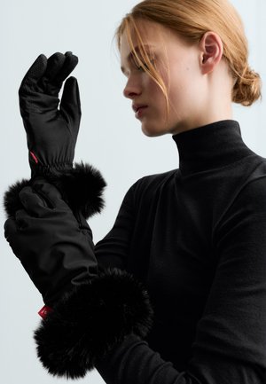 Une femme portant un haut noir ajuste un gant d’hiver noir à large bordure en fourrure, sur un fond uni et clair.