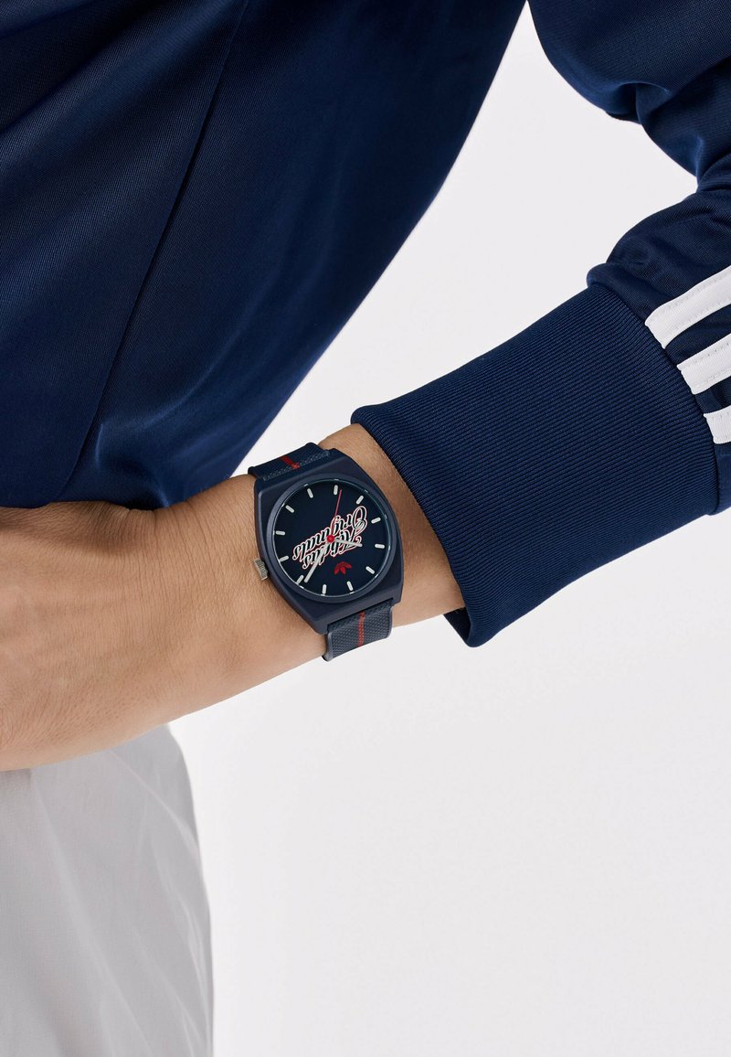 Armbanduhr mit schwarzem Zifferblatt und rotem Schriftzug "Track & Field", getragen am linken Handgelenk über weißen Hosen und einer marineblauen Jacke mit weißen Streifen.