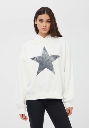 Felpa bianca con un grande motivo a stella grigia in glitter sulla parte anteriore, con cappuccio regolabile e polsini a costine. Sotto, vengono indossati pantaloni neri.