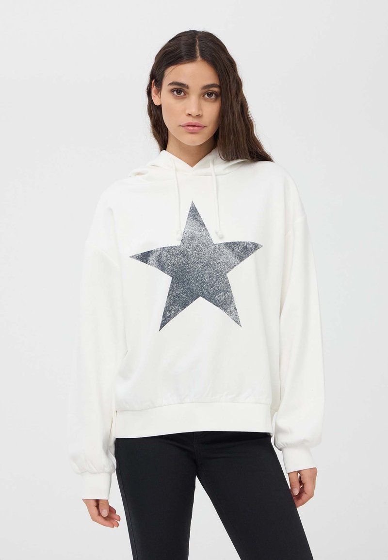 Felpa bianca con un grande motivo a stella grigia in glitter sulla parte anteriore, con cappuccio regolabile e polsini a costine. Sotto, vengono indossati pantaloni neri.