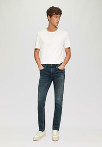 Maglietta bianca a maniche corte abbinata a jeans blu scuri. Design slim-fit con leggero sbiadimento. Indossata con sneakers chiare.