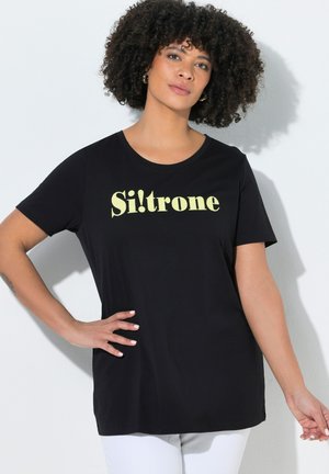 Kvinde med krøllet hår iført sort t-shirt med gul "Si!trone" tekst, poserer med den ene hånd på hoften mod hvid baggrund.