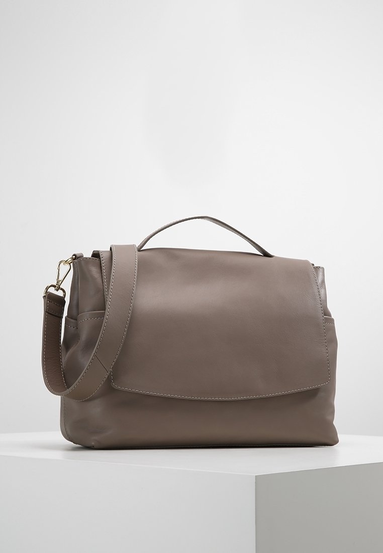 Sac à bandoulière en cuir taupe avec poignée supérieure, fermeture à rabat et sangle réglable, présenté sur une surface blanche avec un fond uni.