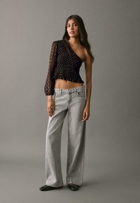 Top nero testurizzato monospalla con orlo arricciato, abbinato a jeans a gamba larga grigio chiaro. La modella indossa scarpe basse nere. Sfondo neutro.