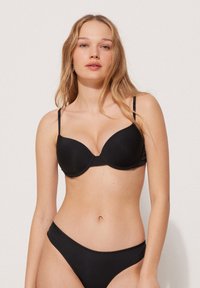 Černá podprsenka s hladkým materiálem, s kosticemi pro podporu a nastavitelnými ramínky. V kombinaci s odpovídajícími černými bikinami.