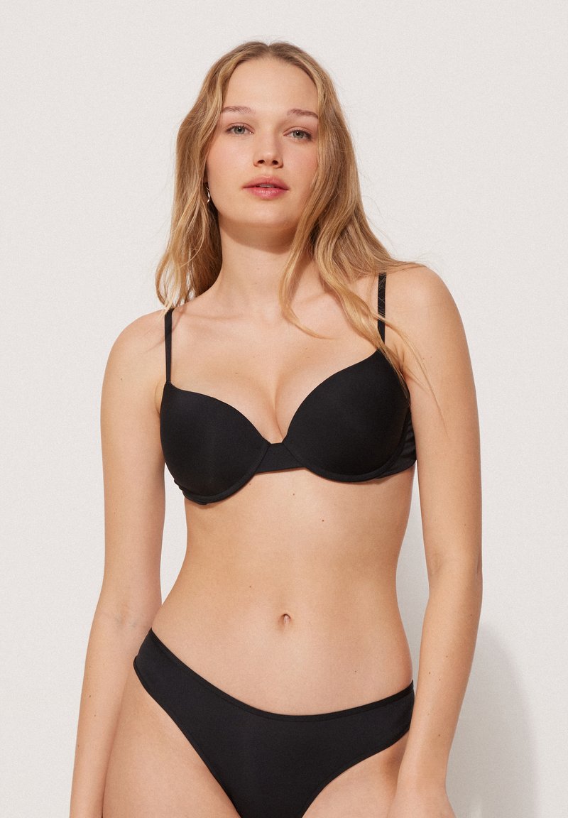 Černá podprsenka s hladkým materiálem, s kosticemi pro podporu a nastavitelnými ramínky. V kombinaci s odpovídajícími černými bikinami.