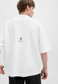 T-shirt blanche surdimensionnée en tissu doux, avec un design graphique noir au dos comportant du texte et un motif circulaire. Manches courtes.