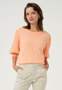 Pull en tricot orange clair avec des manches courtes plissées et un col rond, associé à un pantalon beige. Texture lisse, coupe décontractée.