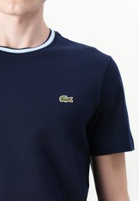 Jūras zils īsās piedurknes T-krekls, kas izgatavots no kokvilnas, ar gaismas zila ribotā apkakli un mazu zaļu izšūtu krokodila logo uz krūtīm.