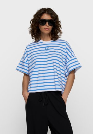 Femme aux cheveux bouclés portant de grandes lunettes de soleil noires, un t-shirt blanc ample à rayures bleues et un pantalon noir avec les mains dans les poches.