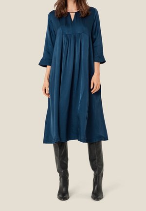 Woman wearing a loose-fitting, knee-length navy blue dress with 3/4 sleeves and black knee-high leather boots, standing on a plain background.