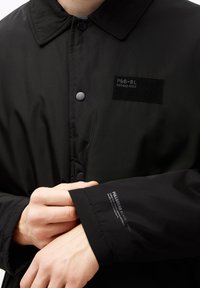 Czarna kurtka z zapięciem na guziki, z naszywką oznaczoną jako "P&G-BL" oraz logo na rękawie. Gładki materiał, minimalna tekstura.