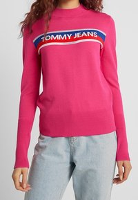 Fuchsia Pullover aus Strickstoff mit hohem Halsausschnitt, gerippten Bündchen und einem horizontalen Logodesign in Blau, Rot und Weiß über der Brust.