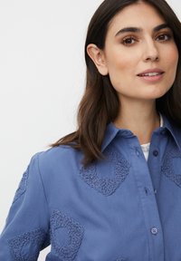 Młoda kobieta z brązowymi włosami nosi niebieską koszulę zapinaną na guziki z fakturowanymi geometrycznymi łatami, delikatnie się uśmiechając na tle białym.