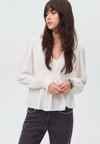 Kvinna med långt mörkt hår iklädd en vit texturerad blus med rynkade ärmar och svarta jeans med nitar på fickorna, som står mot en neutral bakgrund.