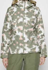 Tarnmuster Softshelljacke in Grün, Weiß und Pink. Mit Reißverschluss, Kapuze mit Kunstfellbesatz und Reißverschlusstaschen an den Seiten.