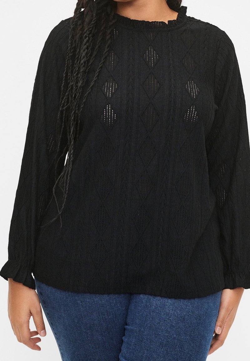 Femme portant un pull en maille noir à manches longues avec des motifs en losange, associé à un jean bleu, montrée des épaules aux hanches.