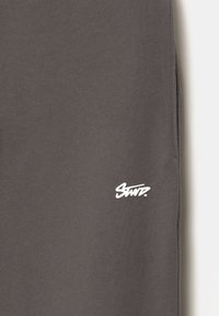 Grå bomuldsstof med en glat tekstur, prydet med et hvidt "Stuz" logo i nederste venstre hjørne. Enkel design, ingen yderligere mønstre.