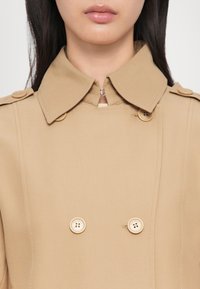 Beiger doppelt geknöpfter Trenchcoat mit spitzem Kragen, zwei Knöpfen und einem Reißverschluss, gefertigt aus einem glatten, leichten Stoff.